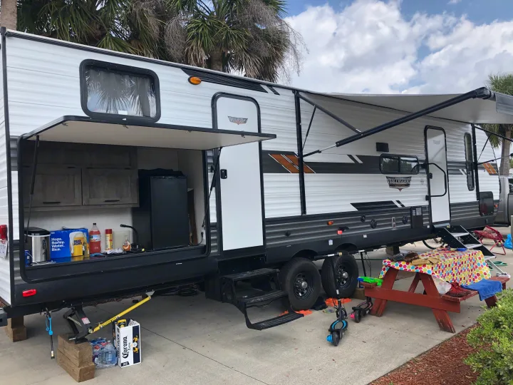 This RV rental, a Forest River Wildwood travel trailer, is set up at a sunny campsite with an outdoor kitchen, awning, and picnic area.. Forest River Wildwood 2020