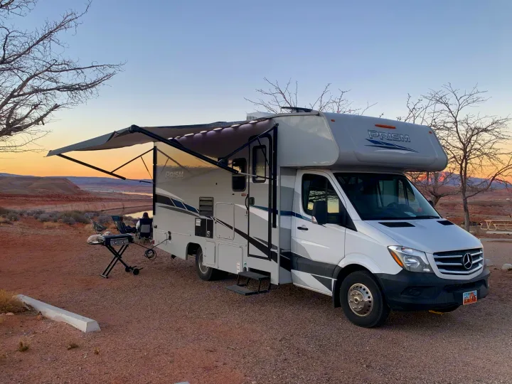 A grey and white Prism RV rental parked at a scenic desert campsite with a lake view. Awning out, grill, and dog at sunset.. Forest River Coachmen Prism 2020
