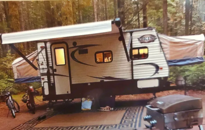 Viking hybrid RV rental ready for camping at a wooded site, complete with extended awning, pop-out beds, bikes, and a grill.. Coachmen Viking 2018