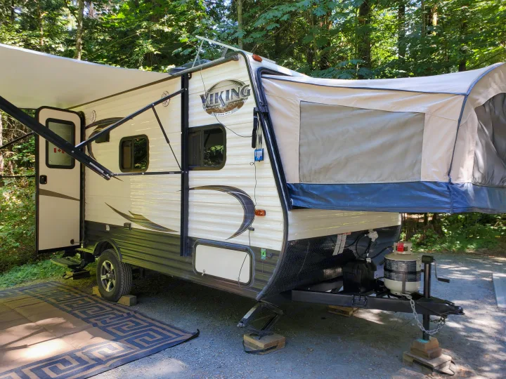 A hybrid RV rental with its awning extended, parked at a sunny campsite amidst green trees, ready for adventure.. Coachmen Viking 2018