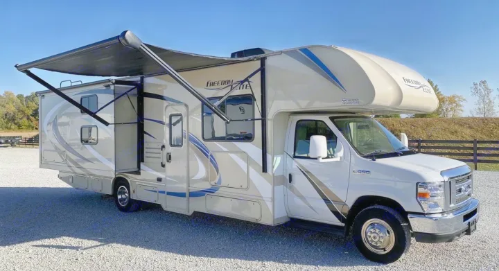 A tan and white Class C Freedom Elite RV rental with its awning extended is parked on a gravel lot under a clear sky.. Thor Motor Coach Freedom Elite 2019