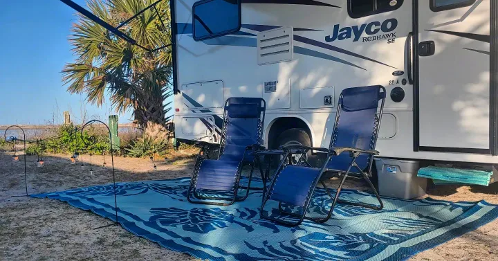 RV rental setup: Jayco RV, chairs, mat, & lights by palm tree and water. Ideal camping relaxation.. Jayco Redhawk 2020