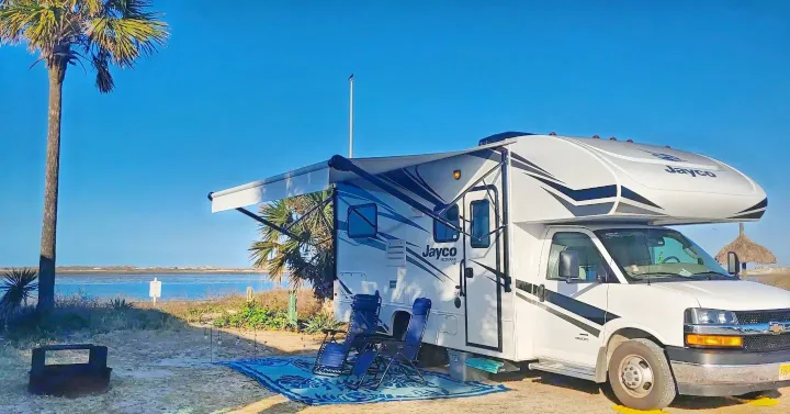 Jayco rv rental parked by the water, perfect for outdoor living. Awning out, chairs set, with serene bay and palm tree views.. Jayco Redhawk 2020