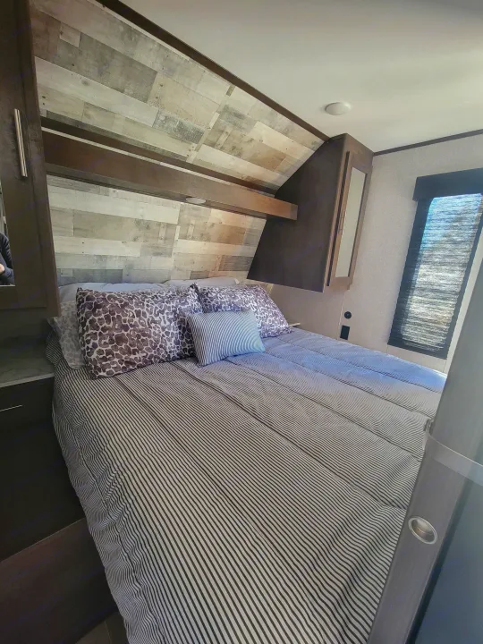 Comfortable RV rental bedroom with striped bedding, decorative pillows, wood plank wall, and overhead storage.. Coleman 263 BH Lantern Edition 2021