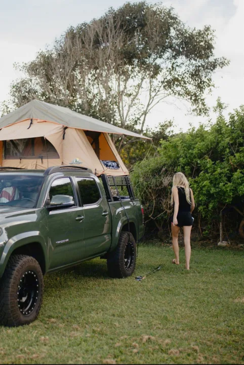 A green pickup truck with a roof-top tent set up for camping. A woman walks nearby. A unique rv rental option for adventurers.. Toyota Tacoma 2021