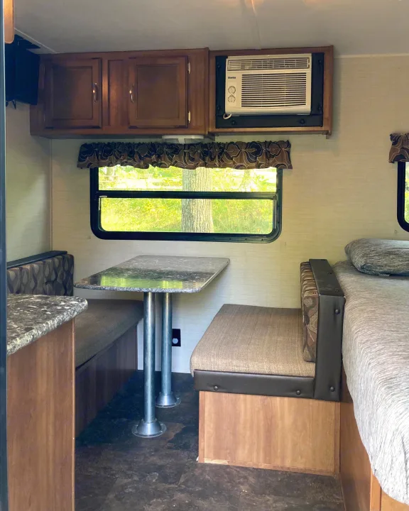 Interior of an RV rental showing a dinette, bed, overhead cabinets, a small TV, and a window AC unit.. Keystone RV Summerland Mini 2016