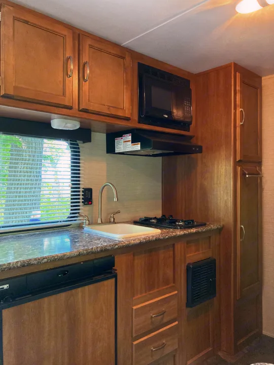 Inside an RV rental, a compact kitchen features wood cabinetry, a sink, gas stove, microwave, and refrigerator.. Keystone RV Summerland Mini 2016