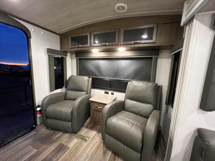 RV rental interior featuring two dark recliners, overhead cabinets, and a doorway with an 'EXIT' sign, showing dusk outside.. Keystone Cougar Half-Ton 2021