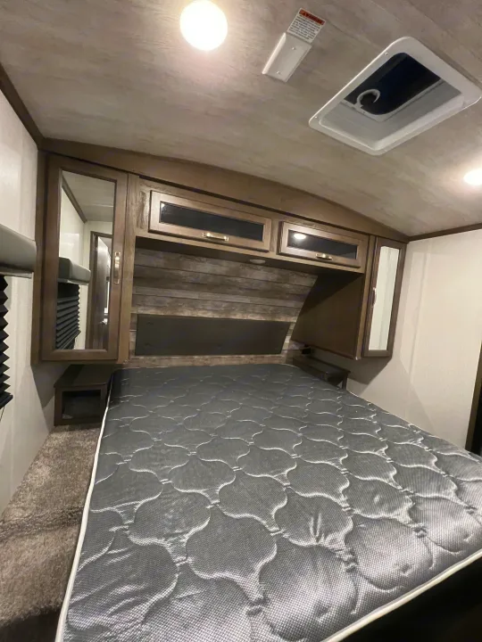 A cozy RV rental bedroom features a grey mattress, wood-paneled headboard, overhead cabinets, and a ceiling vent.. Keystone Cougar Half-Ton 2021