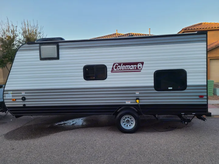 Coleman travel trailer RV rental, parked outdoors. Side view shows windows, connections, and water on the asphalt.. Coleman Latern 17b 2022