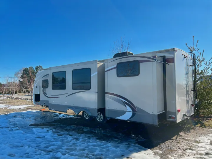 Large RV rental, a beige fifth-wheel travel trailer with slide-outs extended, parked on a snowy lot under a clear sky.. Grand Design Reflection 2015