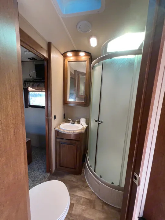 RV rental bathroom features a toilet, sink vanity, curved shower, and skylight. View into a carpeted room with a window.. Grand Design Reflection 2015