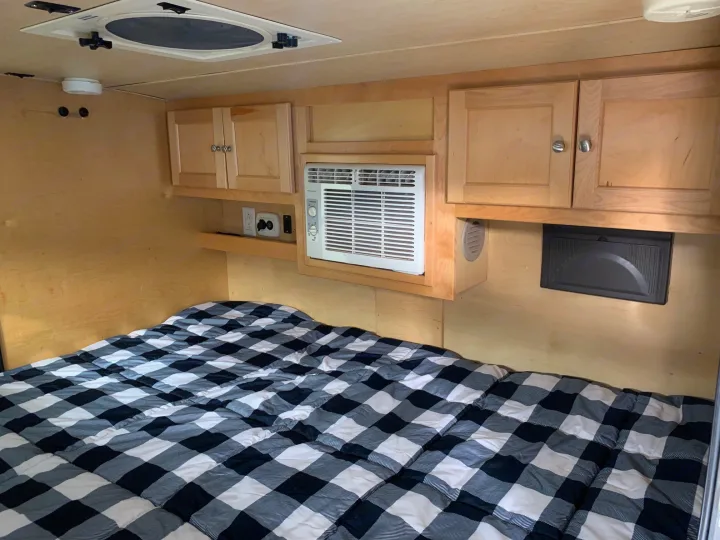 Experience this cozy RV rental bedroom. It features a bed with plaid bedding, overhead cabinets, and a convenient AC unit.. Little Guy Silver Shadow 2015