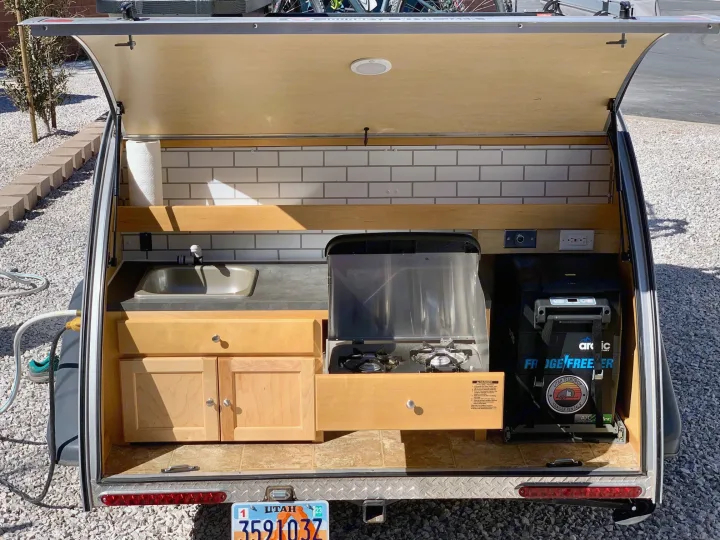 Open rear hatch of a Utah teardrop RV rental, showing its outdoor kitchen with sink, stove, and fridge.. Little Guy Silver Shadow 2015