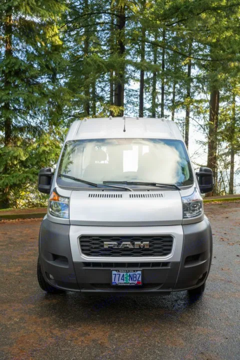 Silver Ram ProMaster van, ideal for RV rental, parked in a forest setting. Front view shows grille, headlights.. RAM ProMaster 2021