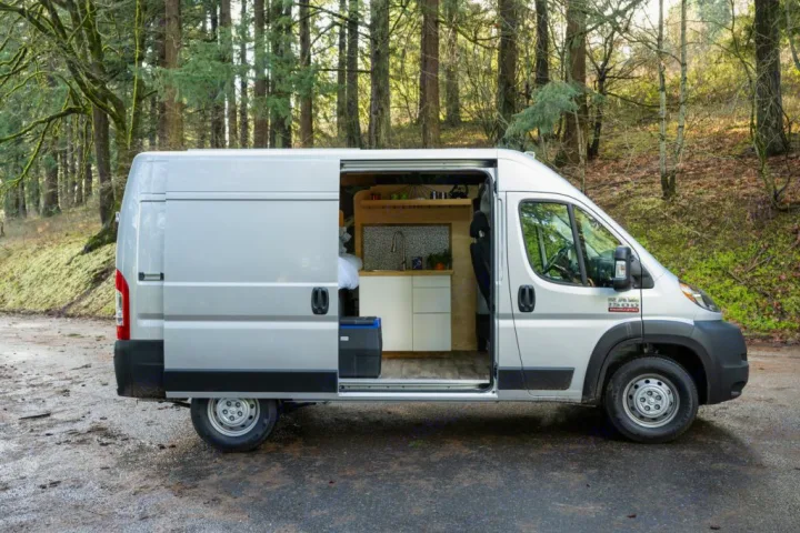 This RV rental shows an open side door revealing a cozy camper van conversion with a kitchen in a forest setting.. RAM ProMaster 2021