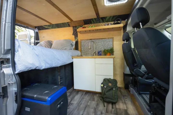 Cozy RV rental interior with bed, kitchen sink, and portable fridge/cooler. Front seats visible, ready for adventure.. RAM ProMaster 2021