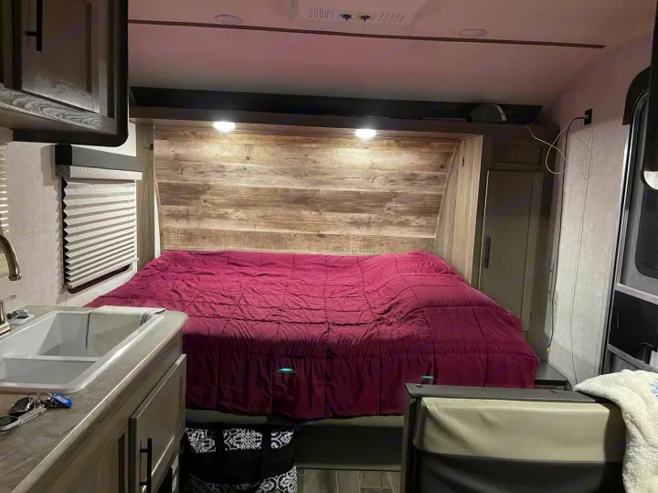 This RV rental features a cozy bedroom with a large purple bed, sink, cabinets, and window blinds, perfect for travel.. Gulf Stream Enlighten 2022