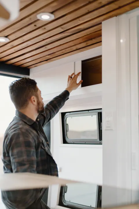A man opens a sliding overhead cabinet in an RV rental, revealing interior storage below a wooden ceiling.. Dodge Sprinter 2019
