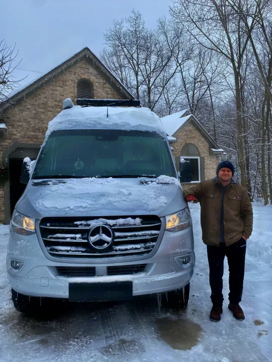 Man smiles next to his snow-covered Mercedes-Benz RV rental van, ready for winter travel adventures.. Thor Sprinter 2022