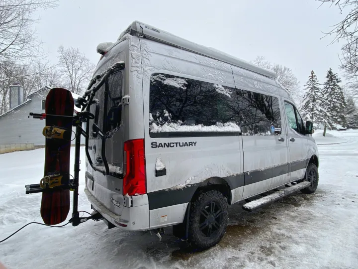 A snow-covered white RV rental camper van, with a snowboard mounted on the back, is parked in a snowy winter driveway.. Thor Sprinter 2022