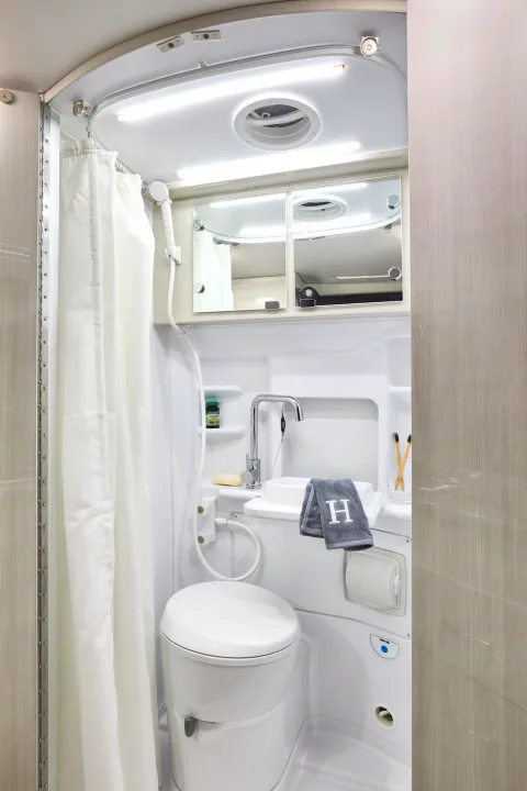 Compact RV rental wet bath. White shower curtain, toilet, sink, mirrored cabinet, and ceiling lights are visible.. Thor Sprinter 2022