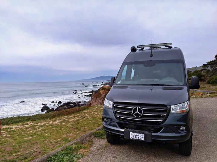 Dark gray Mercedes-Benz camper van (REC VAN) on a cloudy day overlooking a rocky California coast, ideal for an RV rental.. Thor Sanctuary 2022
