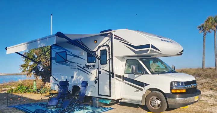 Jayco RV rental parked with awning extended by the calm blue water. Relaxing beach setup with chairs & rug for a sunny getaway.. Jayco Redhawk 2020