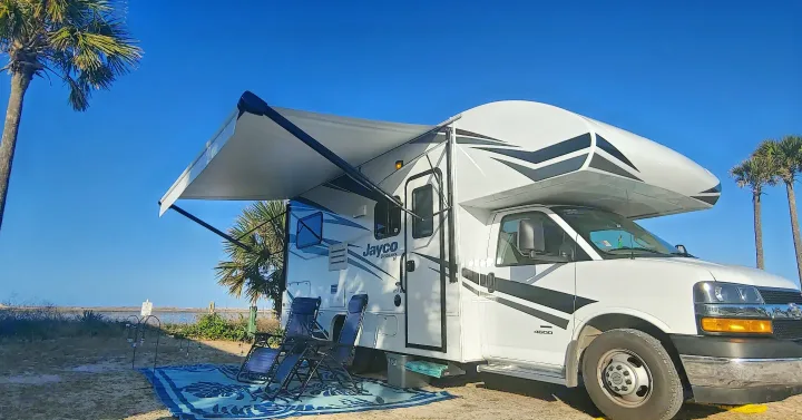 White RV rental with extended awning and outdoor seating on a sandy campsite, offering beachfront views and a clear blue sky.. Jayco Redhawk 2020