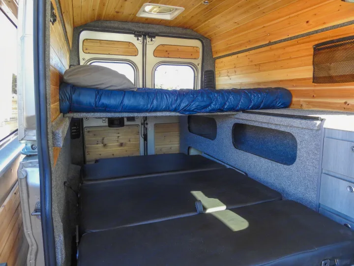 Inside a modern RV rental: a wood-paneled camper van with a cozy bed, lower seating/bed, and smart storage.. Dodge Promaster- Summit X Edition OA9 2021