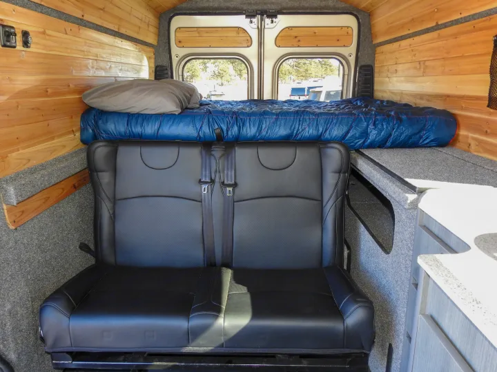 Spacious RV rental interior featuring a comfy bed, two-person seat, and wood paneling, perfect for adventure.. Dodge Promaster- Summit X Edition OA9 2021