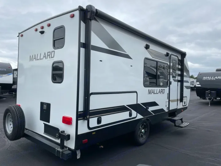 White Mallard travel trailer, ideal for RV rental, parked outdoors. View of back, side, and spare tire.. Heartland Mallard 180BH 2022