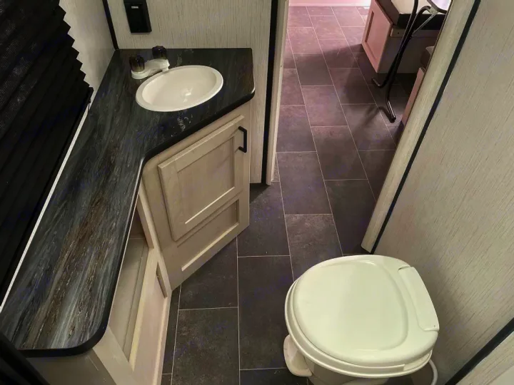RV rental's functional bathroom: vanity with sink, toilet, light wood-grain storage, and dark tiled floor.. Heartland Mallard 180BH 2022