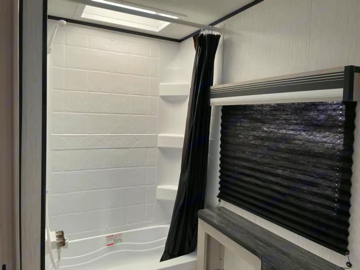 Compact RV rental bathroom with a white shower/tub, black shower curtain, and pleated window blind.. Heartland Mallard 180BH 2022