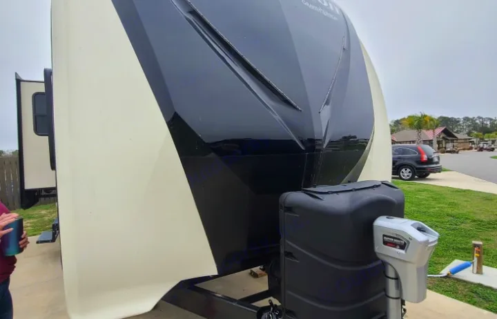 Front view of a large beige & black RV rental, showing its propane tank and electric jack at a campground.. GRAND DESIGN 312BHTS 2019