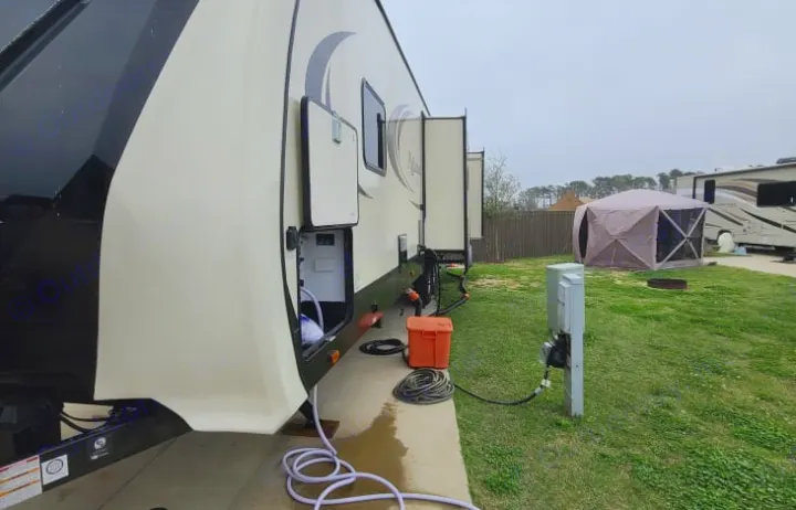 Side view of a large cream and black RV rental, parked on a concrete pad at a campsite. Utility hookups are connected, and slide-outs are extended, ready for use.. GRAND DESIGN 312BHTS 2019