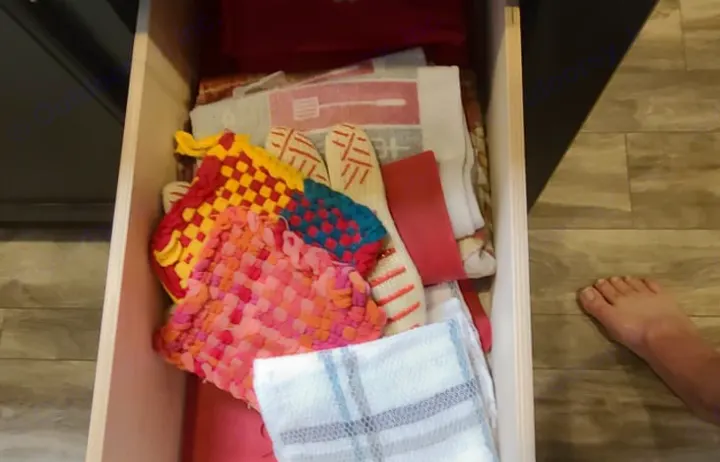 Colorful pot holders & kitchen towels fill a drawer in an RV rental. A bare foot rests on the wood-look floor.. GRAND DESIGN 312BHTS 2019