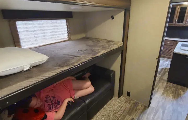 An RV rental interior shows stacked bunk beds; a person relaxes on the lower bunk's couch, kitchen beyond.. GRAND DESIGN 312BHTS 2019