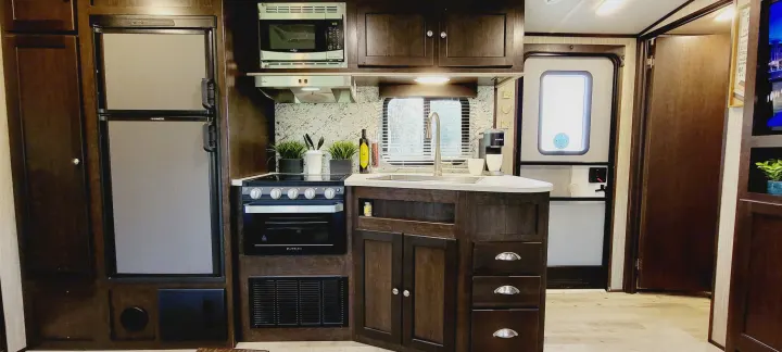Modern RV rental kitchen featuring dark wood cabinets, a fridge, microwave, stove, oven, and sink area.. Heartland RVs Sundance 2018