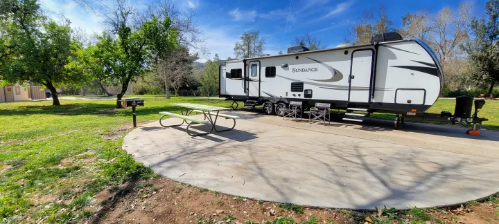 RV rental parked on a concrete pad with a picnic table & grill at a sunny campsite amidst green trees.. Heartland RVs Sundance 2018