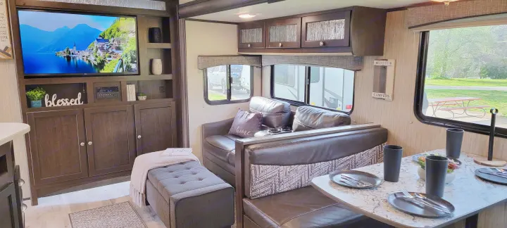 This inviting RV rental interior showcases a cozy living space with a TV, sofa, and ottoman, plus a set dining area, offering comfort on the road.. Heartland RVs Sundance 2018