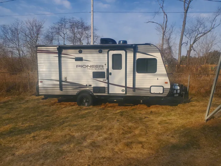 RV rental: White Pioneer travel trailer parked on dry grass; fence & bare trees visible under blue sky.. Heartland RVs Pioneer 2020
