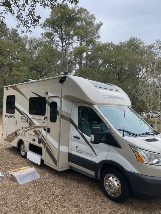 An off-white Compass RV rental, a motorhome, is parked on a gravel campsite amidst trees. Ready for travel.. Thor Compass 2016