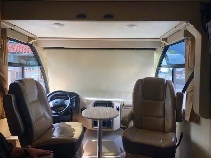 Inside an RV rental, showing the driver's cockpit with two large, tan seats, dashboard, and a small central table.. Thor Vegas 2017