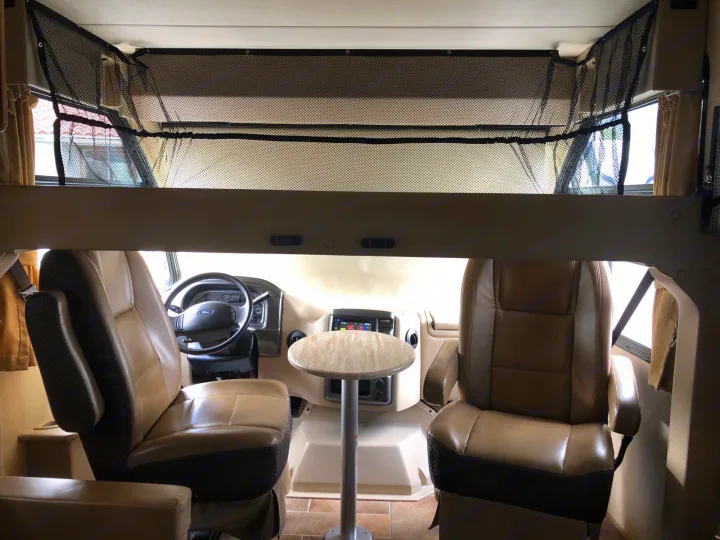 Spacious RV rental front cabin shows tan swivel seats, dashboard, and overhead safety net with a small table.. Thor Vegas 2017