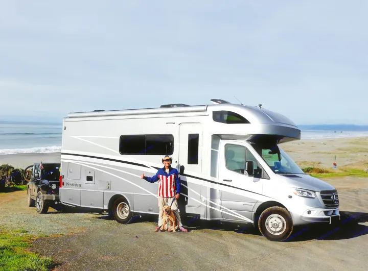 A man and his dog stand by a silver RV rental, towing a black Jeep on a coastal road by the ocean.. Winnebago Navion 2020