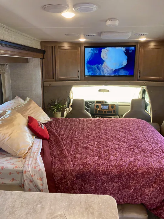 Spacious RV rental bedroom featuring a cozy bed with flamingo sheets, paisley quilt, overhead TV, and a front cab view.. Gulf Stream B Touring Cruiser 2019