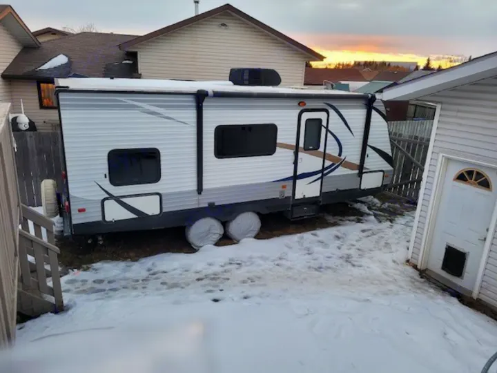 A large travel trailer, often used as an rv rental, is parked in a snowy residential backyard under a twilight sky.. prowler lynx 22LX 2015