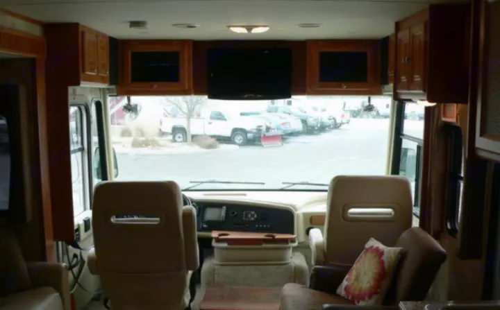Spacious RV rental interior featuring plush captain's chairs, dashboard, overhead TVs, and ample wood cabinetry. Parked view.. Other Other 2010