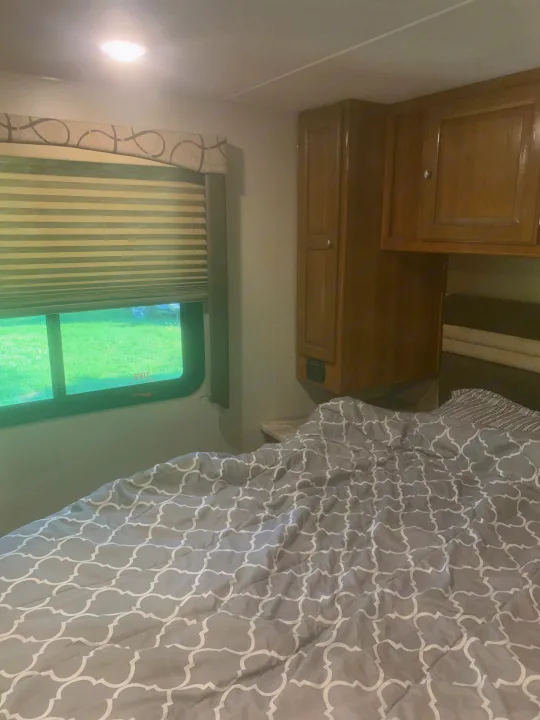 Cozy RV rental bedroom featuring a patterned comforter, window with green view, and wooden storage cabinets.. Forest River Coachmen Leprechaun 2018